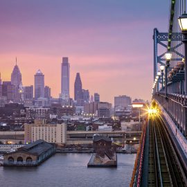 Philadelphia under a hazy purple sunset. An incoming train crosses Ben Franklin Bridge.