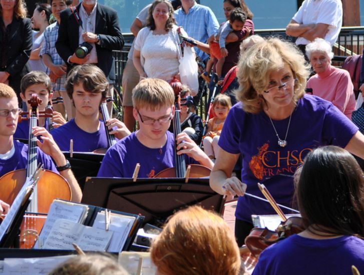 a group of high school students wearing purple shirts play stringed musical instruments in an outdoor concert