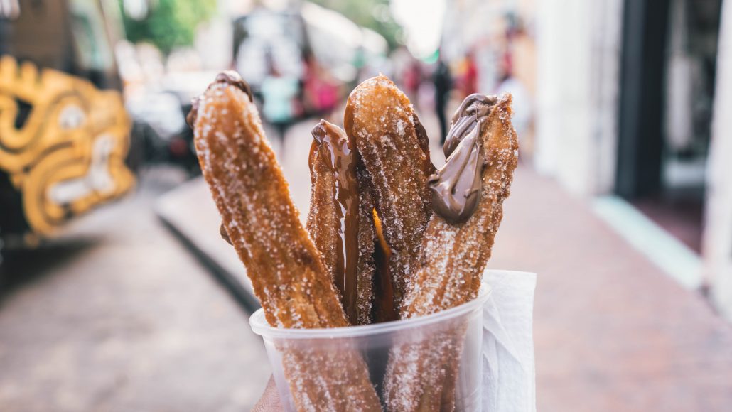 In focus cup filled with churros topped with chocolate, in front of a blurred street background 