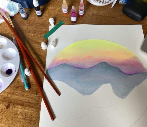 Watercolor painting of mountains and sunset on table with paintbrushes and paint 