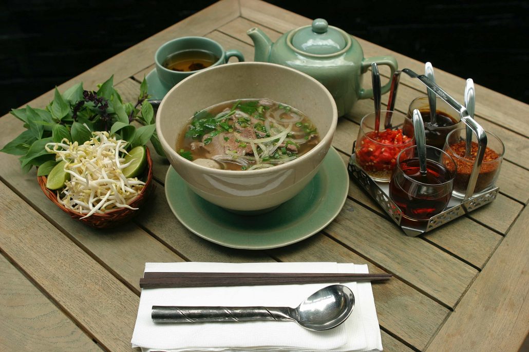 Bowl of Pho with teapot and cup of tea, tray of sauces, and side of bean sprouts, asian basil, and limes