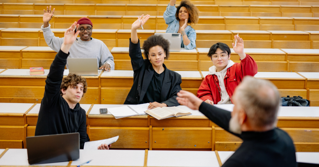 class of five people all raising their hands to answer a question posed by a teacher with a black turtleneck and white beard