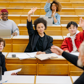 class of five people all raising their hands to answer a question posed by a teacher with a black turtleneck and white beard